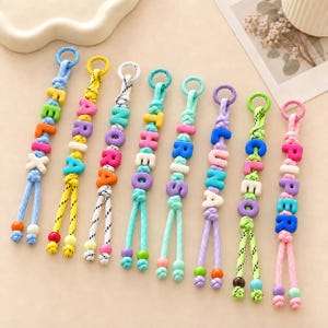 Può includere: Otto portachiavi in corda intrecciata colorata con perline a lettera in plastica che compongono i nomi Felix, Aria, Nora, Theo, Iris, Luna, Cleo e Piper. Ogni portachiavi include un anello circolare in alto e due piccole perline alle estremità.