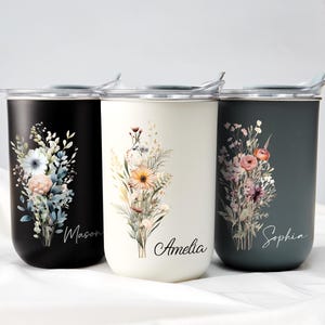 Op de afbeelding: Drie ge&iuml;soleerde reisbekers in zwart, cr&egrave;me en blauwgroen. Elke beker heeft een bloemenboeketontwerp met de namen: Mason, Amelia en Sophia. De bekers hebben zilveren deksels en zijn ontworpen om dranken warm of koud te houden.