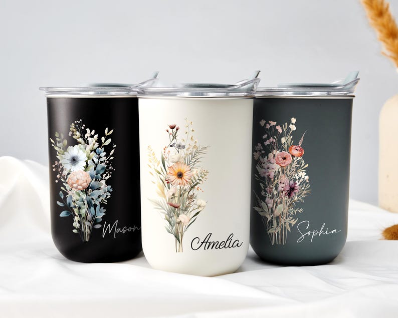 Op de afbeelding: Drie ge&iuml;soleerde reisbekers in zwart, cr&egrave;me en blauwgroen. Elke beker heeft een bloemenboeketontwerp met de namen: Mason, Amelia en Sophia. De bekers hebben zilveren deksels en zijn ontworpen om dranken warm of koud te houden.
