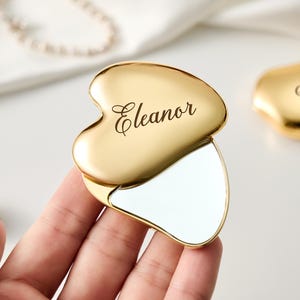May include: A gold-colored compact mirror with the name "Eleanor" engraved on the top. The mirror is heart-shaped and has a reflective surface. Another gold compact with the name "Sophie" is visible in the background.