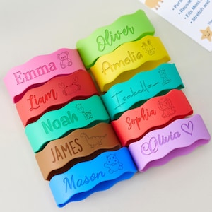 May include: A selection of brightly coloured, wavy silicone bands, each engraved with a name and a small icon. The bands are in various colours, including pink, green, yellow, and blue. The names include "Oliver", "Amelia", "Isabella", "Sophia", "Olivia", "Emma", "Liam", "Noah", "James", and "Mason".