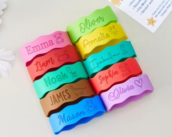 May include: A collection of ten colorful, wavy silicone bands, each engraved with a unique name and a corresponding icon. The names include Emma, Liam, Noah, James, Mason, Oliver, Amelia, Isabella, Sophia, and Olivia, featuring icons like a bear, crab, giraffe, dinosaur, hippo, rabbit, and heart.