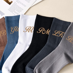 Può includere: Sei paia di calzini a coste in vari colori: marrone, blu, bianco, nero, blu navy e grigio. Ogni calzino presenta iniziali ricamate in oro. I calzini sono disposti ordinatamente, mostrando le diverse opzioni di colore e i dettagli personalizzati.