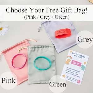 May include: Three satin drawstring gift bags in pink, grey, and green, each paired with a silicone bottle band. The grey bag features an orange band with the name Olivia. A white informational card displays the text Bottle Labels Name Bands.