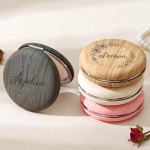Peut inclure: Plusieurs miroirs compacts personnalisés. L'un est ouvert, révélant le miroir, avec "Sophia" gravé sur la surface en bois gris foncé. Les autres miroirs sont empilés, avec un grain de bois clair, crème et rose, avec "Aeison" et des détails floraux.