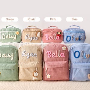 May include: Four corduroy backpacks and four matching lunch bags in green, khaki, pink, and blue. Each item features a personalized name in raised, textured lettering and a small decorative plush accent, such as a flower, lion, or bear.
