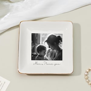 May include: Square white ceramic trinket dish with a gold rim. The center features a black and white photo of a mother and child. The words "Mom, I love you" are written below the photo. A pearl bracelet is in the background.