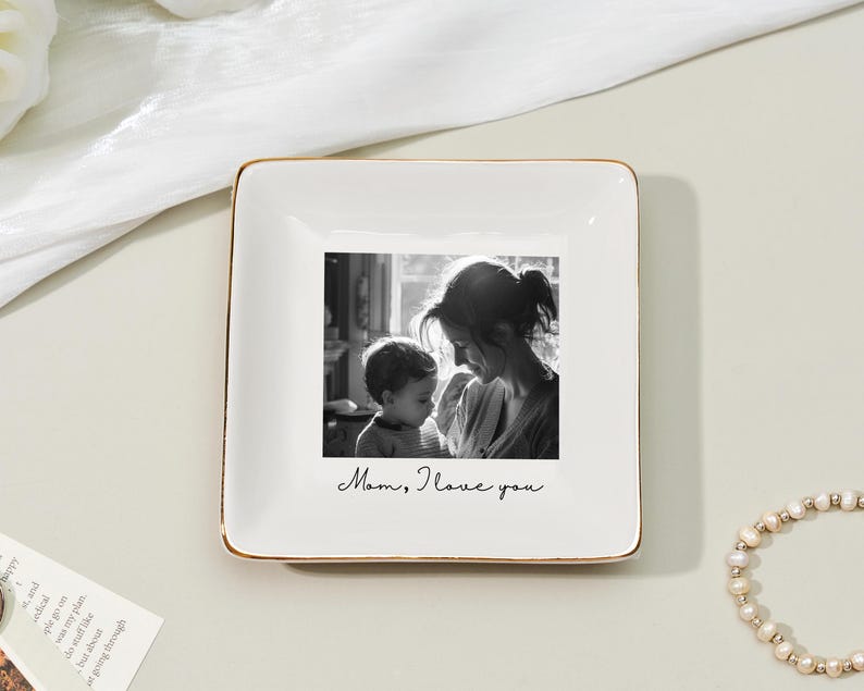 May include: Square white ceramic trinket dish with a gold rim. The center features a black and white photo of a mother and child. The words "Mom, I love you" are written below the photo. A pearl bracelet is in the background.