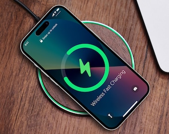 May include: A silver smartphone rests on a wireless charging pad, displaying a green lightning bolt icon indicating fast charging. The phone screen shows a gradient of green and purple hues. The charging pad has a green ring and is connected to a black cable.