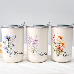 Op de afbeelding: Drie cr&egrave;mekleurige ge&iuml;soleerde bekers met bloemmotieven en namen. Elke beker heeft een ander aquarelboeket: blauw en paars, lavendel en oranje en rood. De namen "Mason", "Amelia" en "Grace" zijn in script geschreven.