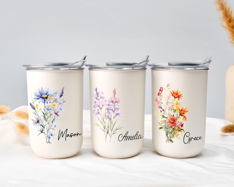 Op de afbeelding: Drie cr&egrave;mekleurige ge&iuml;soleerde bekers met bloemmotieven en namen. Elke beker heeft een ander aquarelboeket: blauw en paars, lavendel en oranje en rood. De namen "Mason", "Amelia" en "Grace" zijn in script geschreven.