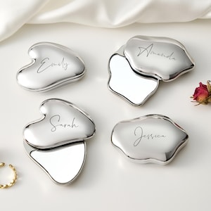 May include: Four silver compact mirrors with personalized names: Emily, Amanda, Sarah, and Jessica. The compacts have a unique shape and open to reveal a mirror. The background includes dried roses and a gold ring.
