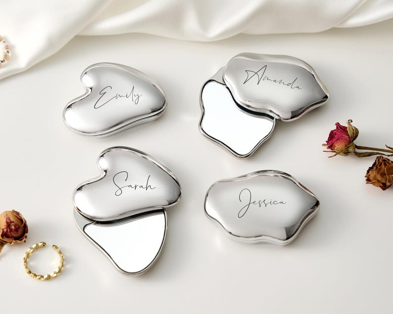 May include: Four silver compact mirrors with personalized names: Emily, Amanda, Sarah, and Jessica. The compacts have a unique shape and open to reveal a mirror. The background includes dried roses and a gold ring.