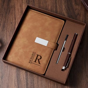 Könnte beinhalten: Ein braunes Lederjournal mit silbernem Verschluss, einem Stift und einem Stylus, präsentiert in einer Geschenkbox. Das Journal ist mit dem Namen "RICHARD" und dem Buchstaben "R" geprägt. Stift und Stylus sind silber und braun.