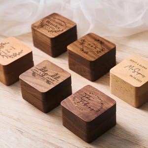 May include: A collection of six square wooden boxes, each with a hinged lid. The boxes are engraved with names, dates, and floral designs. The wood tones vary from light to dark brown, and the boxes are arranged on a light wood surface.