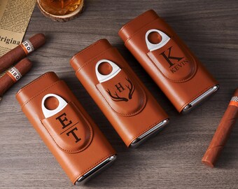 May include: Three brown leather cigar cases with silver accents and built-in cigar cutters. Each case features a personalized design: initials "E T", a deer antler graphic, and the name "KEVIN". Cigars are visible on the wooden surface.