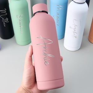 May include: A pink insulated water bottle with the name "Archie" in silver script. Other bottles in various colors, each with a different name, are visible in the background. The bottles have a matte finish and a black cap.