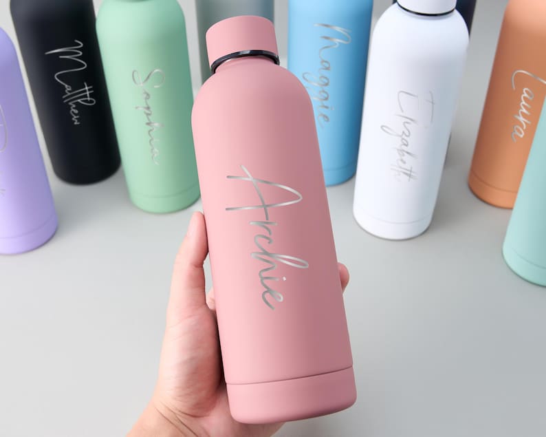 May include: A pink insulated water bottle with the name "Archie" in silver script. Other bottles in various colors, each with a different name, are visible in the background. The bottles have a matte finish and a black cap.