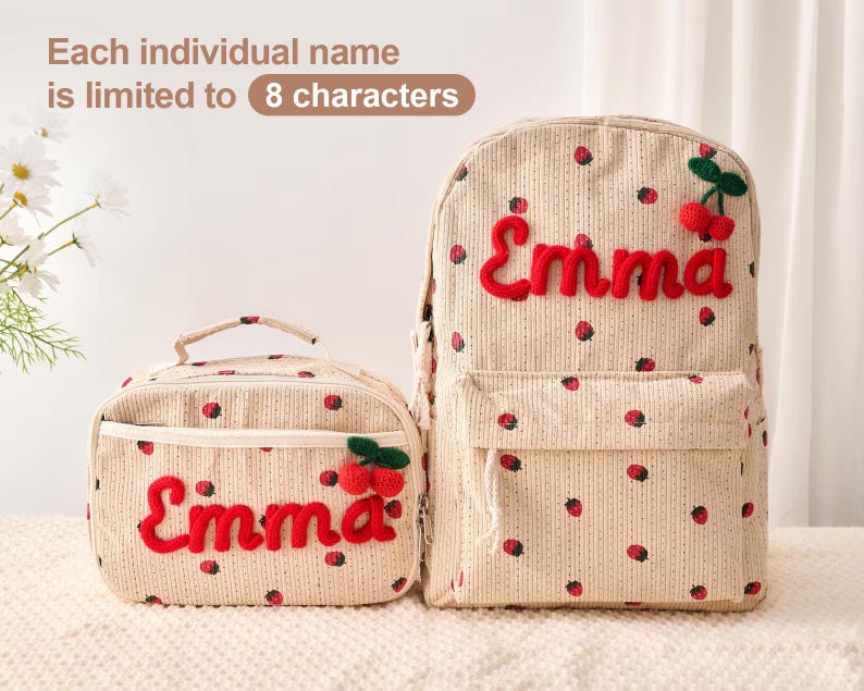 May include: A beige corduroy backpack and matching lunch bag featuring a red strawberry pattern. Both items display the name Emma in red embroidered script with a small red cherry applique. Text above states: Each individual name is limited to 8 characters.