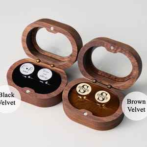 May include: Two wooden cufflink boxes with oval-shaped lids are open, revealing cufflinks. One box has black velvet, the other brown. The cufflinks have personalized text and initials. The boxes are made of dark wood.