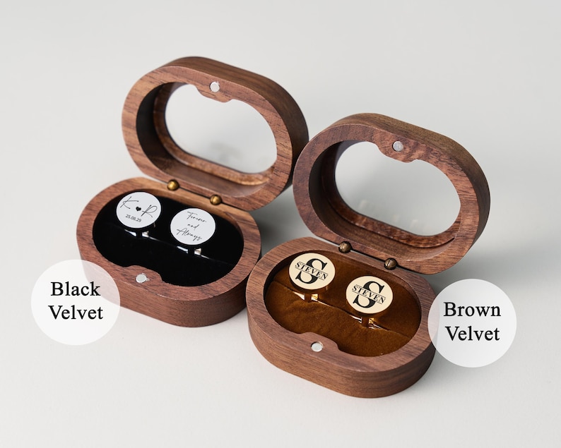 May include: Two wooden cufflink boxes with oval-shaped lids are open, revealing cufflinks. One box has black velvet, the other brown. The cufflinks have personalized text and initials. The boxes are made of dark wood.