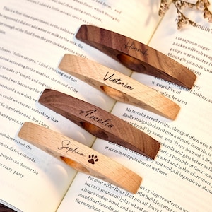 May include: Four wooden book page holders with personalized names: Hannah, Victoria, Amelia, and Sophia. Two are light-colored wood, and two are dark. The Sophia holder has a paw print. The holders are resting on an open book.