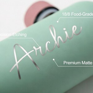 May include: A light green water bottle with the name "Archie" etched in silver. The bottle has a premium matte finish and is made of 18/8 food-grade steel. The bottle's lid is pink.