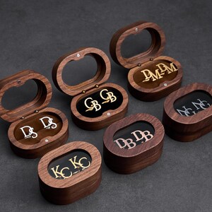 May include: Several wooden jewelry boxes with oval-shaped lids are open, revealing personalized cufflinks. The cufflinks feature initials in silver and gold tones. The boxes are arranged on a dark gray surface.