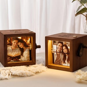Peut inclure: Deux boîtes carrées en bois brun foncé avec un éclairage interne chaud et une manivelle latérale. Chaque boîte affiche une photographie d'un couple. Le haut de la boîte gauche est gravé Cheers to Love, et la droite Our Story Begins.