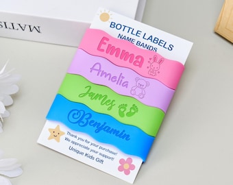 May include: Four silicone bottle labels or name bands in pink, purple, lime green, and blue, each featuring a name and icon: Emma with a rabbit, Amelia with a teddy bear, James with footprints, and Benjamin. They are displayed on a white card.