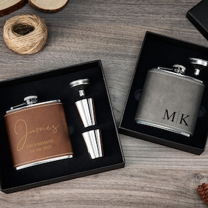 Könnte beinhalten: Zwei Hüftflaschen in schwarzen Boxen. Eine ist braun mit dem Namen "James" und "Groomsman 04-08-2023" in goldener Schrift. Die andere ist grau mit den Initialen "MK" in Schwarz. Beide haben silberne Deckel und werden mit einem Trichter und zwei Schnapsgläsern geliefert.
