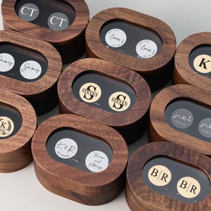 May include: A collection of wooden boxes with oval-shaped lids, each containing a pair of personalized cufflinks. The cufflinks feature initials, names, and dates on circular backgrounds. The boxes are made of dark wood and have a clear window to display the cufflinks.