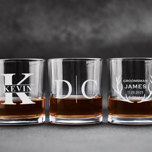 Può includere: Tre bicchieri da whisky trasparenti riempiti di liquido ambrato. Ogni bicchiere è personalizzato con testo bianco. Un bicchiere ha la lettera "K" e il nome "KEVIN", un altro le lettere "D" e "C", e il terzo dice "GROOMSMAN JAMES 11.20.2025".