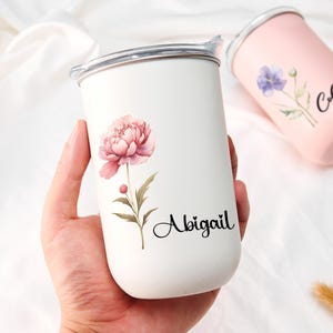 Op de afbeelding: Een witte beker met een roze pioenroos en de naam "Abigail" erop gedrukt. Een tweede roze beker met een paarse bloem en de naam "Collen" is ook zichtbaar. Beide bekers hebben een zilveren rand en een deksel.