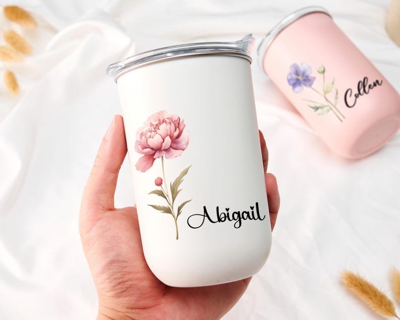Op de afbeelding: Een witte beker met een roze pioenroos en de naam "Abigail" erop gedrukt. Een tweede roze beker met een paarse bloem en de naam "Collen" is ook zichtbaar. Beide bekers hebben een zilveren rand en een deksel.