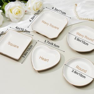 May include: A collection of white ceramic dishes in various shapes: square, wavy square, heart, round, and wavy round. Each dish has a gold rim and the shape name printed in gold. Measurements are provided in inches and centimeters.