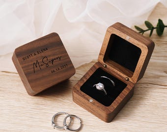 May include: A wooden ring box with a hinged lid. The box is engraved with the text "SCOTT & ELENA McSopers 06.16.2025". The box is open and a ring is inside.