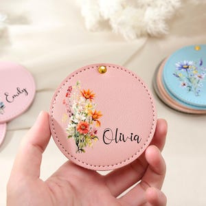 Peut inclure: Un miroir de poche rond rose avec un motif floral et le nom "Olivia" imprimé en script noir. Le miroir a un clou doré. D'autres miroirs de poche avec des motifs floraux et des noms sont visibles.