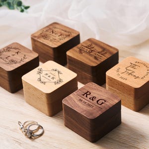 May include: Six wooden ring boxes with engraved personalized messages. The boxes are made of light and dark wood and have a square shape. The boxes are arranged in a semi-circle on a light wood surface.