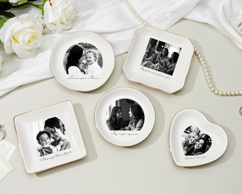 May include: A collection of white ceramic trinket dishes in various shapes: round, square, and heart-shaped. Each dish features a black and white photograph and a gold rim. The dishes are inscribed with phrases like "Love you Mom" and "My forever friends."