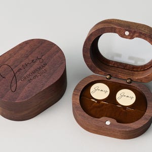May include: A wooden cufflink box with a clear lid, open to reveal two gold cufflinks. The box is engraved with the name "James" and the word "Groomsman" along with a date. The box is oval-shaped and made of dark wood.