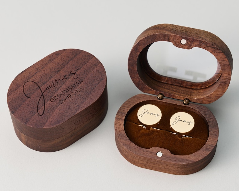 May include: A wooden cufflink box with a clear lid, open to reveal two gold cufflinks. The box is engraved with the name "James" and the word "Groomsman" along with a date. The box is oval-shaped and made of dark wood.