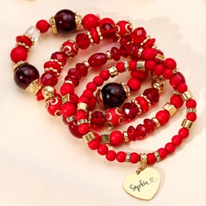 May include: Four red beaded bracelets with gold accents and a heart-shaped charm. The bracelets feature various bead sizes and shapes, including round, faceted, and square beads. One charm is engraved with the name "Sophia".