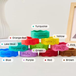 May include: A collection of eleven colorful silicone wristbands stacked in a pyramid. Each band features a unique name and icon, including Benjamin, Ella, Sophia, Harper, Olivia, James, Amelia, Oliver, Lucas, and others in turquoise, orange-red, yellow, lake blue, green, pink, blue, purple, red, and brown.