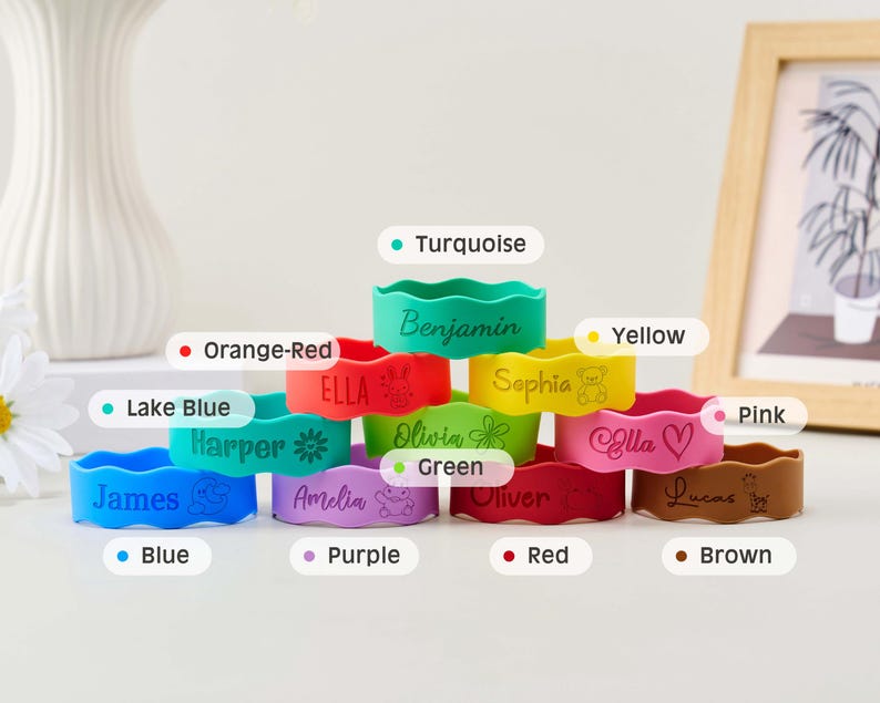 May include: A collection of eleven colorful silicone wristbands stacked in a pyramid. Each band features a unique name and icon, including Benjamin, Ella, Sophia, Harper, Olivia, James, Amelia, Oliver, Lucas, and others in turquoise, orange-red, yellow, lake blue, green, pink, blue, purple, red, and brown.
