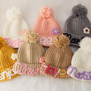 Könnte beinhalten: Sieben gestrickte Mützen mit Bommeln und bestickten Namen. Die Mützen sind in verschiedenen Farben, darunter Weiß, Rosa, Grau, Gelb und Braun. Jede Mütze hat eine kleine Blumenverzierung.