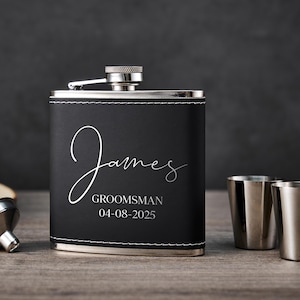 Op de afbeelding: Zwarte flacon met zilveren accenten en de naam "James" in witte letters. De flacon is gelabeld met "Groomsman" en een datum. Twee zilveren borrelglaasjes en een zilveren trechter zijn ook zichtbaar.