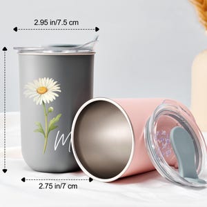 Op de afbeelding: Twee roestvrijstalen reisbekers. E&eacute;n is grijs met een madeliefjesafbeelding en het woord "m" in het wit. De andere is roze, met een doorzichtig deksel. De grijze beker is 12 cm hoog en heeft een diameter van 7 cm. De roze beker heeft een diameter van 7,5 cm.