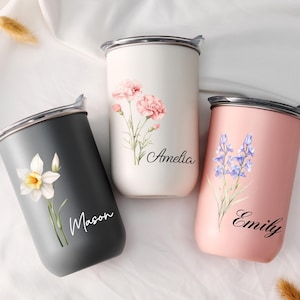 Op de afbeelding: Drie gepersonaliseerde roestvrijstalen bekers met bloemmotieven. E&eacute;n is donkergrijs met een witte narcis en de naam "Mason". Een andere is wit met roze anjers en de naam "Amelia". De derde is roze met blauwe bloemen en de naam "Emily".