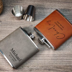 May include: Two personalised hip flasks, one grey and one brown, with silver detailing, are displayed on a wooden surface. The grey flask is engraved with "Michael, Groomsman, 05-12-2025." The brown flask is engraved with "Charley, Groomsman, 04-08-2025." Two small stainless steel shot glasses are also present.